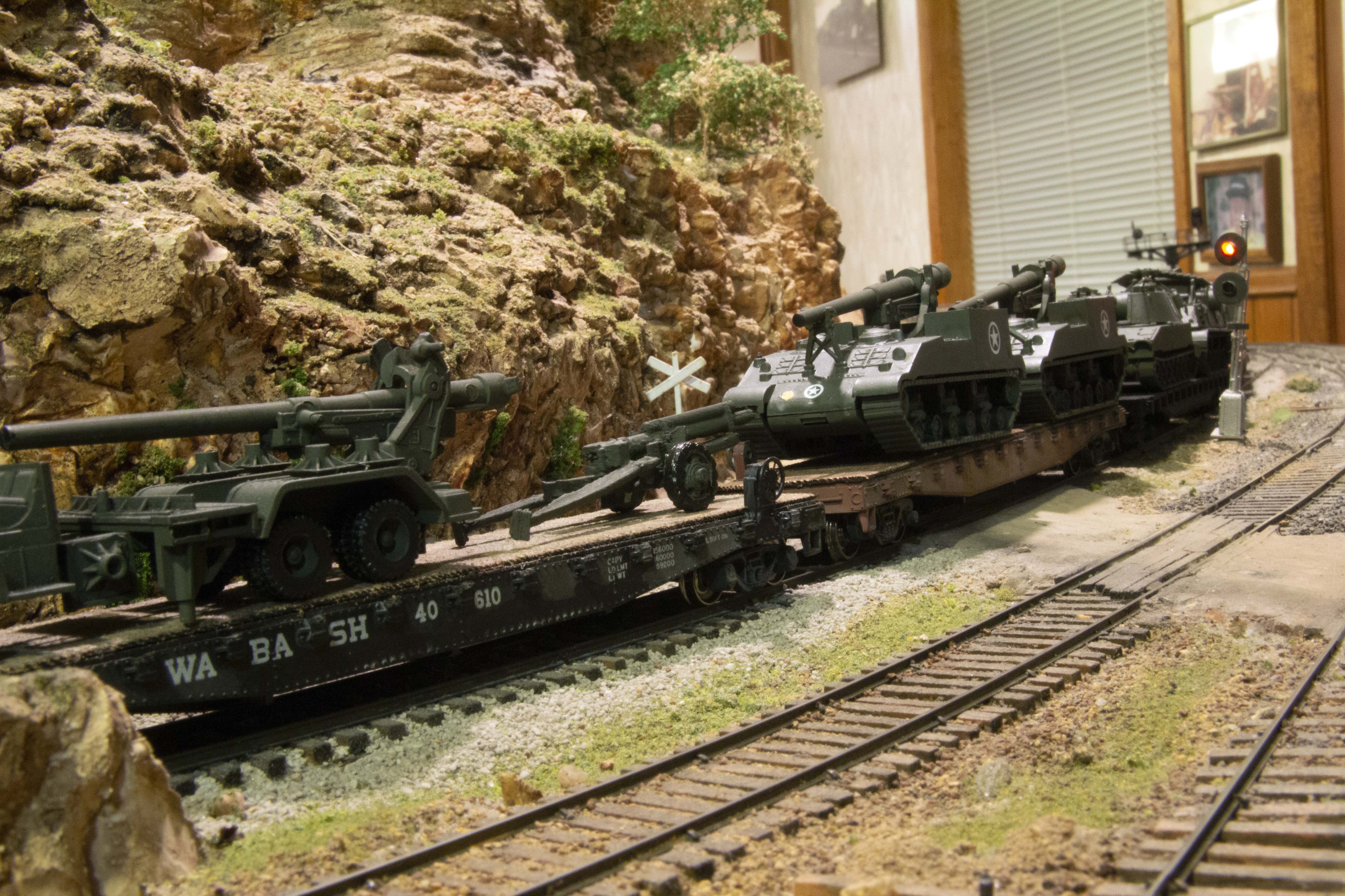 Close up view of 3D-printed military equipment on flatcars passing a signal on the Stoney Creek & Western layout